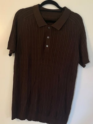 Brun kabelstickad pike - Säljer en brun polotopp med korta ärmar och ribbad struktur. Toppen har klassisk krage och tre knappar framtill. Perfekt till jeans eller kjol för en enkel men snygg look.