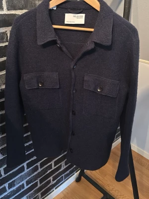 Mörkblå overshirt från Selected Homme - Stilren mörkblå overshirt från Selected Homme med klassisk krage, knäppning framtill och två stora bröstfickor med lock och knapp. Tillverkad i mjukt, stickat material som ger en avslappnad men ändå snygg look. Perfekt att bära över en t-shirt eller skjorta. Skriv gärna om frågor uppstår!🤗