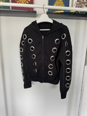 Svart hoodie med stora metallöljetter - Säljer en svart hoodie med dragkedja och stora, runda metallöljetter över hela framsidan och ärmarna. Matchande byxor säljs separat på min profil! Gå gärna in och titta om det är något som låter intressant!