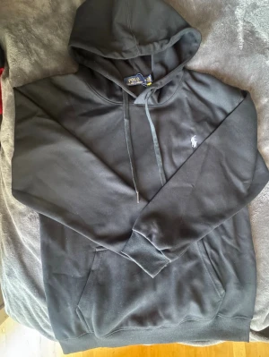 Svart hoodie från Polo Ralph Lauren - Svart hoodie från Polo Ralph Lauren med klassisk huva och dragsnören. Tröjan har en stor magficka och den ikoniska vita Polo-loggan broderad på bröstet. Tillverkad i mjukt material och har ribbade muddar vid ärmslut och nederkant.