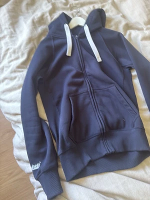Russemerch - Mörkblå hoodie med vit dragsko och dragkedja framtill. Två fickor på magen och ribbade muddar vid ärmslut och nederkant. Märket Dress broderat i vitt på ena ärmen. Perfekt för en chill och avslappnad stil.