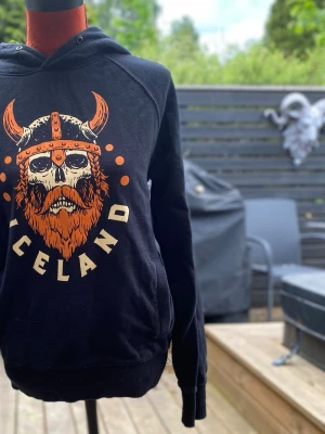 Svart hoodie med vikingatryck Iceland S - Svart huvtröja med ett stort tryck på framsidan föreställande en vikingaskalle med skägg och horn, samt texten "ICELAND" undertill. Tröjan har långa ärmar och en huva. 2 fickor Längd: 65 cm Bystvidd: 49 cm 100% bomull 
