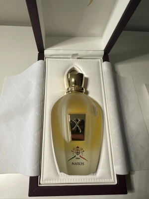 Xerjoff Naxos Eau de Parfum 100ml - Lyxig parfym från Xerjoff, modellen Naxos, i en elegant frostad glasflaska med gulddetaljer och exklusiv guldfärgad kork. Flaskan ligger i en snygg presentask med Xerjoff-logga och italienska flaggans färger på etiketten. Volym: 100 ml.