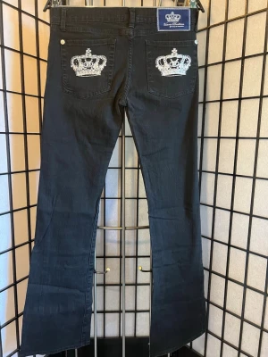 Victoria Beckham for Rock & Republic Jeans – Y2K Bootcut / McBling 👑✨ - Ikoniska Y2K-jeans från Victoria Beckham for Rock & Republic. Svart denim med de klassiska kronorna broderade på bakfickorna, dekorerade med strassdetaljer. Snygg bootcut-passform som ger den där tidiga 2000-talslooken som är hetare än någonsin.  Perfekta för Y2K, McBling, Paris Hilton- och celeb-core-stilen.  📏 Mått: • Midja: ca 37–38 cm plant • Innerben: ca 81 cm • Totallängd: ca 104 cm • Lårvidd: ca 25 cm • Fotvidd: ca 21 