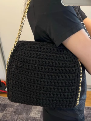 Handgjord svart virkad axelväska med guldkedja  - Handgjord svart virkad axelväska i mycket fint skick, endast använd ett fåtal gånger. Väskan har en stilren design med guldfärgad kedjerem och magnetknäppning som håller innehållet säkert på plats.
