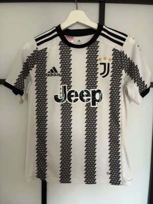 Juventus matchtröja Adidas svartvit - Juventus officiella matchtröja från Adidas i svartvitt med grafiskt mönster och Jeep-logga på bröstet. Tröjan har korta ärmar, rund hals och tre svarta ränder på axlarna. Tillverkad i lätt och ventilerande material, perfekt för fotboll eller träning.