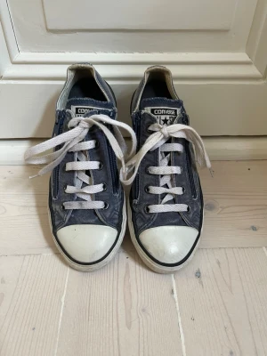 Blå-grå Converse All Star låga sneakers med dragkjedja på båda sidor - Klassiska låga Converse All Star sneakers i mörkblå canvas med vita gummidetaljer, vita snören och dragkjedja på båda sidor. Ikonisk logga på plösen och tåhätta i gummi. Perfekta för en avslappnad och cool stil.