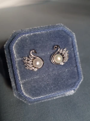 Eternal Love (silver 925 stift)  - Svanar är en symbol för kärlek, trohet och evighet – de representerar starka band och livslång samhörighet. Smycket är dekorerat med en sötvattenspärla, CZ-diamanter som reflekterar ljuset och ger en exklusiv lyster.                                    Material: Rhodium, Stift i 925 silver (stämplat), sötvattenspärlor, CZ-diamant Storlek: längd ca 1,5 cm Bredd ca 1,5 cm -Inga prisförslag, tack                                        - Varan packas noggrant och säkert 