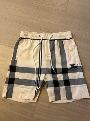 Beige rutiga shorts från Burberry - Snygga beige shorts från Burberry med klassiskt rutigt mönster i svart och grått. Shortsen har elastisk midja med vit snörning och Burberry-logga på benet. Perfekta för sommaren och ger en lyxig känsla till din outfit. Små i storlek!