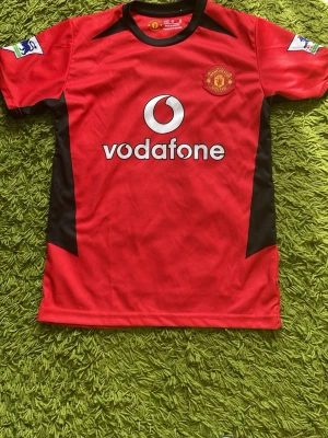 Manchester United Beckham tröja - Klassisk röd Manchester United matchtröja med svarta detaljer, Vodafone-logga på bröstet och klubbmärke. På ryggen står det 'Beckham' och nummer 7. Korta ärmar och tillverkad i lätt, ventilerande material. Perfekt för fotbollsfans och samlare.