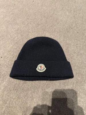 Mörk blå/ marin ribbstickad mössa från Moncler - Svart ribbstickad mössa från Moncler med klassisk uppvikt kant och broderad logotyp framtill. Perfekt för kalla dagar och enkel att matcha med olika outfits. Snygg och stilren design som funkar året runt. Daily wear (pris kan diskuteras)