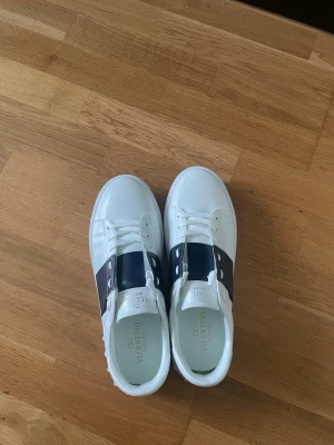 Valentino Garavani  - Snygga vita sneakers från Valentino Garavani med bred mörkblå remsa över mitten och klassiska vita snören. Skorna har en låg profil och är tillverkade i slätt läder med subtila nitar på sidorna för en extra edge.