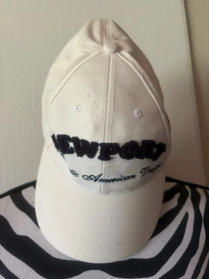 Vit Newport keps med brodyr - Snygg vit keps från Newport med broderad text framtill och bak. Framtill står det 'Newport' och baktill 'Rhode Island, USA'. Justerbar rem bak och klassisk böjd skärm. Tillverkad i bomullstyg, perfekt för en clean och stilren look.