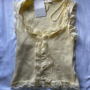 Gul ribbad spetsdetaljerad topp Zara - Supersöt ljusgul ribbad topp från Zara med spetsdetaljer längs kanterna och knappar framtill. Ärmlös modell med volangkant vid axlar och nederkant. Perfekt för en fräsch och feminin look.