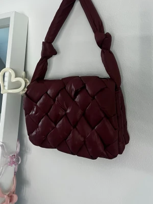 Mörkröd vadderad handväska - Säljer en mörkröd handväska med vadderad, flätad design och en mjuk och puffig look.