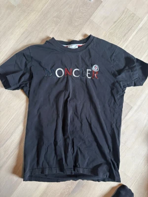 Svart Moncler t-shirt med logga - Svart t-shirt från Moncler med stor broderad logga i vitt och rött på bröstet samt Moncler-märke på vänster sida. Klassisk passform och rund halsringning. Tillverkad i mjuk bomull som känns skön mot huden.