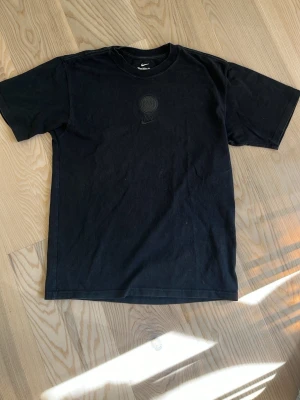Svart loose fit t-shirt från Nike - Svart t-shirt från Nike med diskret logga på bröstet. Modellen är loose fit och har klassisk rund hals och korta ärmar. Perfekt för en avslappnad och sportig stil. Med Paris Saint German logga