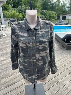 Klassisk camo overshirt - Snygg overshirt i militärgrönt camomönster med tryckknappar framtill och två bröstfickor. Skjortan har axelklaffar och långa ärmar med knapp vid ärmslut. Perfekt lager på lager-plagg för en avslappnad streetstil.