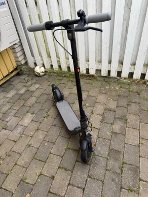 Segway Ninebot Kickscooter F - Säljer en svart elscooter med gråa handtag och bred ståplatta. Scootern har stora hjul och enkel design, perfekt för att ta sig runt i stan. Materialet är mestadels metall och plast. Laddningsuttag och bromshandtag finns på styret.