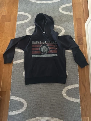 Mörkblå Saint Laurent hoodie - Snygg mörkblå hoodie från Saint Laurent med stor huva och känguruficka. Framsidan har tryck med texten 'SAINT LAURENT' och grafiska ränder i rött och grått. Tillverkad i mjukt bomullsmaterial, perfekt för en avslappnad streetwear-look.