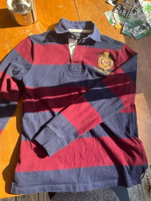 Vintage Ralph lauren rugby shirt - Hej, jag säljer just nu denna extremt snygga polo tröjan.✌️  Den har ett snyggt randig mönster med färgerna blå och röd (se sista bild för bättre vinkel i ljus)  Den har en fin broderad logga på bröstet med välgjorda detaljer 💯  Blå krage med vita knappar💯  Inga defekter✅  Priset kan självklart diskuteras👊