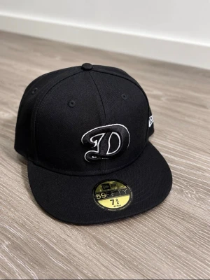 Svart New Era 59Fifty keps - Säljer en svart New Era 59Fifty keps med broderad vit D-logga framtill. Klassisk flat brim och strukturerad passform. Märke på sidan och officiell storleksdekal kvar på skärmen. Perfekt för dig som gillar streetwear och sportig stil.