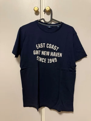 Gant t-shirt - Snygg mörkblå t-shirt från Gant med vit text på bröstet: 'East Coast Gant New Haven Since 1949'. Klassisk rund hals och korta ärmar. Perfekt för en avslappnad och stilren look.