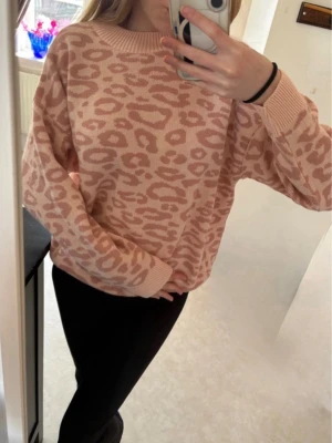 Rosa leopardmönstrad stickad tröja - Säljer en snygg stickad tröja i ljusrosa med leopardmönster i en mörkare rosa nyans. Tröjan har rund halsringning, långa ärmar och ribbade muddar vid ärmslut och nederkant. Perfekt för dig som gillar trendiga mönster och mjuka material.