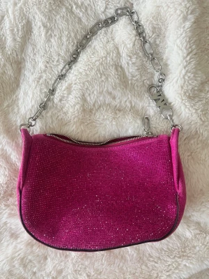 Rosa glittrig handväska från Michael Kors - Säljer en snygg rosa handväska från Michael Kors med glittrande yta och silverfärgad kedja som axelrem. Väskan har dragkedja upptill och ett ljust innerfoder med logga. Perfekt för dig som vill sticka ut med en färgglad accessoar. Går att bära i olika former som visas på sista bilden. 