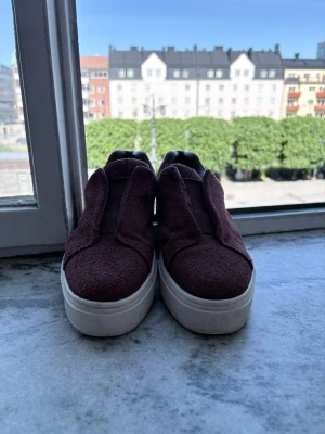Vinröda slip-on sneakers mocka - Säljer ett par vinröda fin skor i mocka med tjock vit sula. Skorna har minimalistisk design utan snörning och elastiska paneler på sidorna för enkel påtagning. Perfekta för dig som gillar stilrena och bekväma sneakers.
