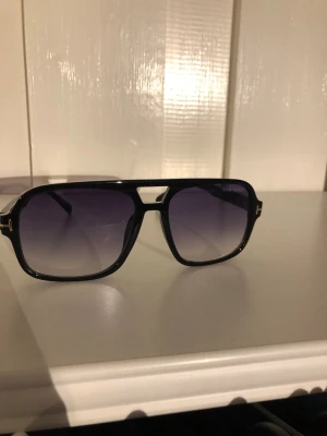 Svarta Tom Ford solglasögon - Snygga svarta solglasögon från Tom Ford med fyrkantig form och mörka, tonade glas. Bågarna har gulddetaljer vid sidorna och logga på linsen. Perfekt accessoar för en trendig look.