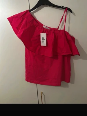 Rosa oneshoulder topp från Ellos - Trendig rosa oneshoulder topp från Ellos med volangdetalj över axeln och smalt axelband på ena sidan. Köpte begangnad men den var för stor så säljer nu, den är mer rosa i verkligheten💕💕 prislapp på