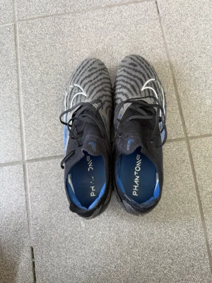 Nike Phantom GT2 grå/svarta fotbollsskor - Snygga Nike Phantom GT2 fotbollsskor i grått och svart med blå detaljer och vita swoosh-loggor. Skorna har ett mönstrat stickat material för bra bollkontroll, snörning och blå yttersula med dobbar för optimalt grepp på planen. Perfekta för dig som vill sticka ut på fotbollsplanen.