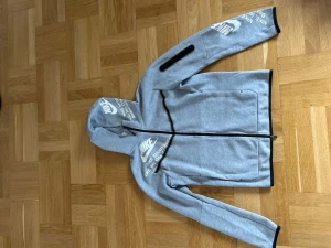 Grå Nike tech hoodie - Snygg grå hoodie från Nike med vit logga och texttryck på bröstet, huvan och ena ärmen. Dragkedja framtill, fickor på sidorna och svart kantband vid ärmslut och nederkant. Perfekt för en sportig och avslappnad stil. Har matchande byxor i min profil:)
