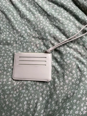 Ljusgrå minimalistisk kortplånbok - Snygg och stilren ljusgrå kortplånbok med tre kortfack på framsidan och ett större fack på baksidan. Plånboken har dragkedja upptill och en avtagbar handledsrem. Tillverkad i slätt konstläder som ger en modern vibe.