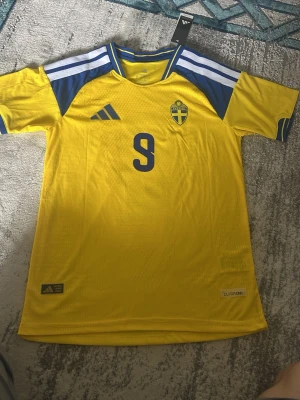 Svensk landslagströja Adidas gul - Gul svensk landslagströja från Adidas med blå detaljer och nummer 9 på bröstet. Tröjan har blått och vitt mönster på axlarna, Sveriges landslagsemblem på vänster sida och är tillverkad i lätt, ventilerande Climalite-material.