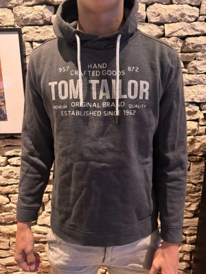 Mörkgrå hoodie från Tom Tailor - Mörkgrå hoodie från Tom Tailor med vit textprint på bröstet och vita dragsnören i huvan. Klassisk passform med långa ärmar och mjukt material, perfekt för en avslappnad stil. Snygg och enkel design som funkar till det mesta.