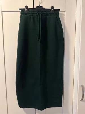Mörkgrön långkjol med snörning - Mysig mörkgrön långkjol med resår i midjan och snörning framtill. Kjolen har praktiska fickor på sidorna och är gjord i ett mjukt sweatshirtliknande material, perfekt för en avslappnad stil.