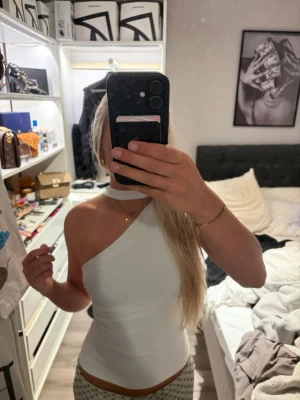 Vit oneshoulder topp med choker-detalj - Snygg vit topp i oneshoulder-modell med en trendig choker runt halsen. Toppens passform är tight och den är ärmlös på ena sidan, vilket ger en cool och modern look. Perfekt för dig som vill sticka ut lite extra.