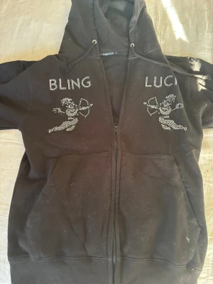 Svart hoodie med bling och ängelmotiv - Svart ziphoodie från BLINGLUCK med huva och dragsko. På bröstet finns texten 'BLING' och 'LUCK' i glittrande stenar samt två änglar med pilbåge i samma bling-stil. Hoodien har fickor framtill och är perfekt för en cool och avslappnad look.