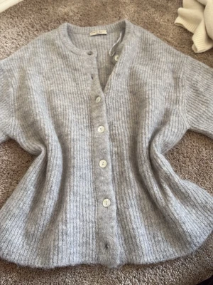 Ljusgrå ribbstickad kofta - Mysig ljusgrå ribbstickad kofta med rund halsringning och stora vita knappar framtill. Koftan har en avslappnad passform och är tillverkad i ett mjukt, fluffigt material som känns lyxigt. Perfekt att slänga över en t-shirt eller topp.
