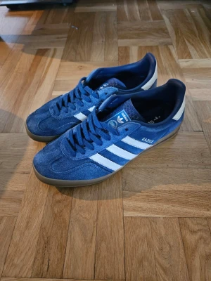 Adidas Gazelle Indoor blå sneakers - Säljer ett par klassiska Adidas Gazelle sneakers i blå mocka med vita detaljer och de ikoniska tre ränderna på sidan. Skorna har gummisula och låg profil, perfekt för en avslappnad och stilren look. Tidlös modell som funkar till det mesta.