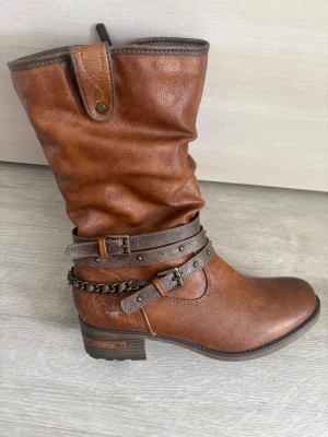 Bruna cowboy boots från Mustang - Snygga bruna cowboy boots från Mustang i konstläder med dekorativa remmar, nitar och kedjedetalj runt ankeln. Bootsens skaft är något rynkat och har en låg klack. Mustang-logga på sulan och präglad häst på sidan.