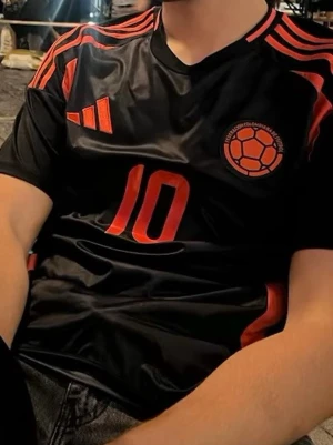 Colombia fotbollströja Adidas svart/röd 2022 - Snygg svart Colombia fotbollströja från Adidas med röda detaljer och nummer 10 på både fram- och baksidan. På ryggen står det 'James' för James Rodriguez och 'Colombia'. Tröjan har korta ärmar och är tillverkad i ett lätt, ventilerande material perfekt för träning eller match.