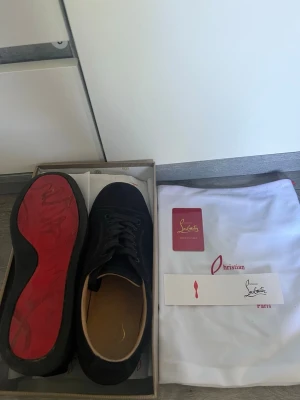 Svarta sneakers från Christian Louboutin - Säljer ett par svarta sneakers från Christian Louboutin med ikoniska röda sulor och diskret logga på sidan. Skorna har en stilren design i läder och textil, med svarta snören och detaljer. Perfekta för dig som vill ha något exklusivt och trendigt.