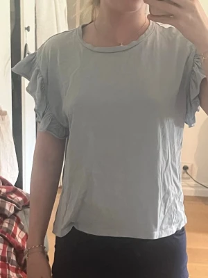 Ljusblå topp med volangärm från Zara - Supersöt ljusblå topp från Zara med volangdetaljer på ärmarna. T-shirten har rund halsringning och är tillverkad i mjukt bomullsmaterial. Perfekt för dig som gillar en enkel men ändå trendig look.