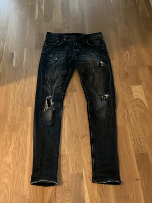 Mörkblå slitna jeans från Nudie storlek W30 L33 - Säljer ett par mörkblå jeans från Nudie med slimfit och slitningar på framsidan. Jeansen har klassiska fem fickor, låg midja och coola detaljer med trasiga partier och Nudie-logga på bakfickorna. Perfekta för en avslappnad streetstil.