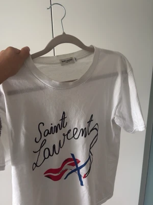 Saint Laurent T-shirt - Säljer nu min Vita Saint Laurent t-shirt med ett snyggt tryck framtill. Anledningen till att jag säljer T-shirten är då jag har växt ur den. Inga defekter och i mycket bra skick.