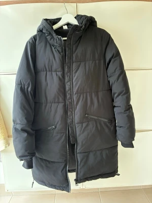 Svart vadderad jacka stl 34 Nyskick - Svart vadderad jacka med huva och dragkedja framtill. Klassisk rak passform. Jackan har två fickor med dragkedja i midjehöjd och en dragsko i nederkant. Ärmsluten har muddar. Slutar strax ovanför knät. H&M Divided. Stl 34, passar en small bra. Använd en gång, så nästan i nyskick.