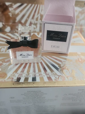 Miss Dior Essence parfym 5 ml från Dior - Miss Dior Essence parfym 5ml från Dior i en elegant glasflaska med ljusrosa doftvätska. Flaskan har en dekorativ svart rosett runt halsen och ett genomskinligt fyrkantigt lock. Förpackningen är ljusrosa med svart etikett och klassisk Dior-design.